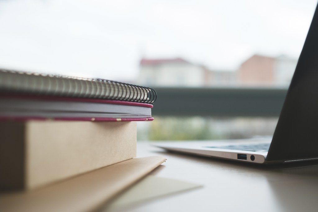 Decorative image of a a stack of books with a spiral bound notebook on top and a laptop computer on the right to illustrate study
